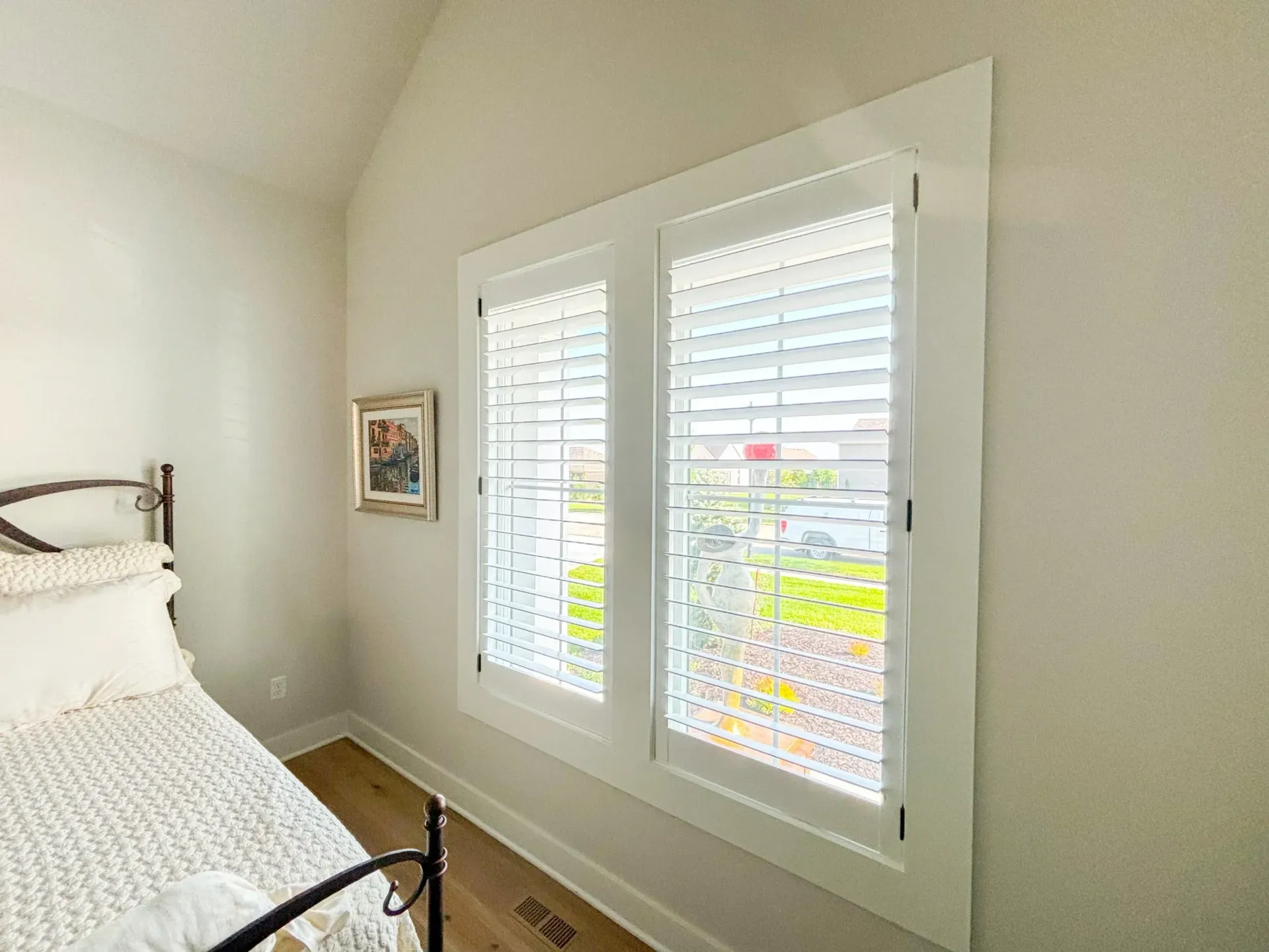 Bedroom with white shutters and a bed, featuring wood floors and a framed artwork on the wall.