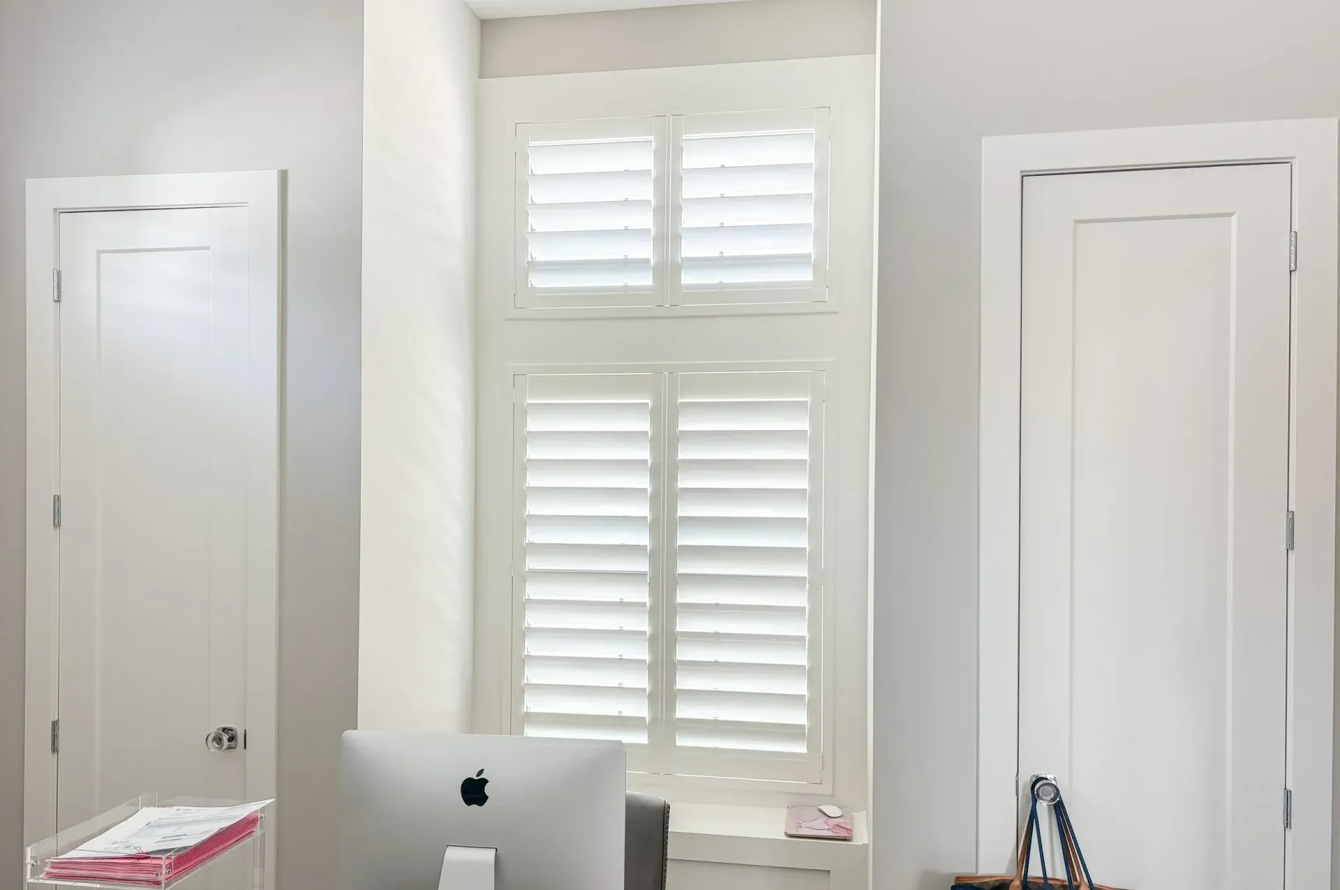White shutters cover a window between two white doors, a desk with a monitor sits below.