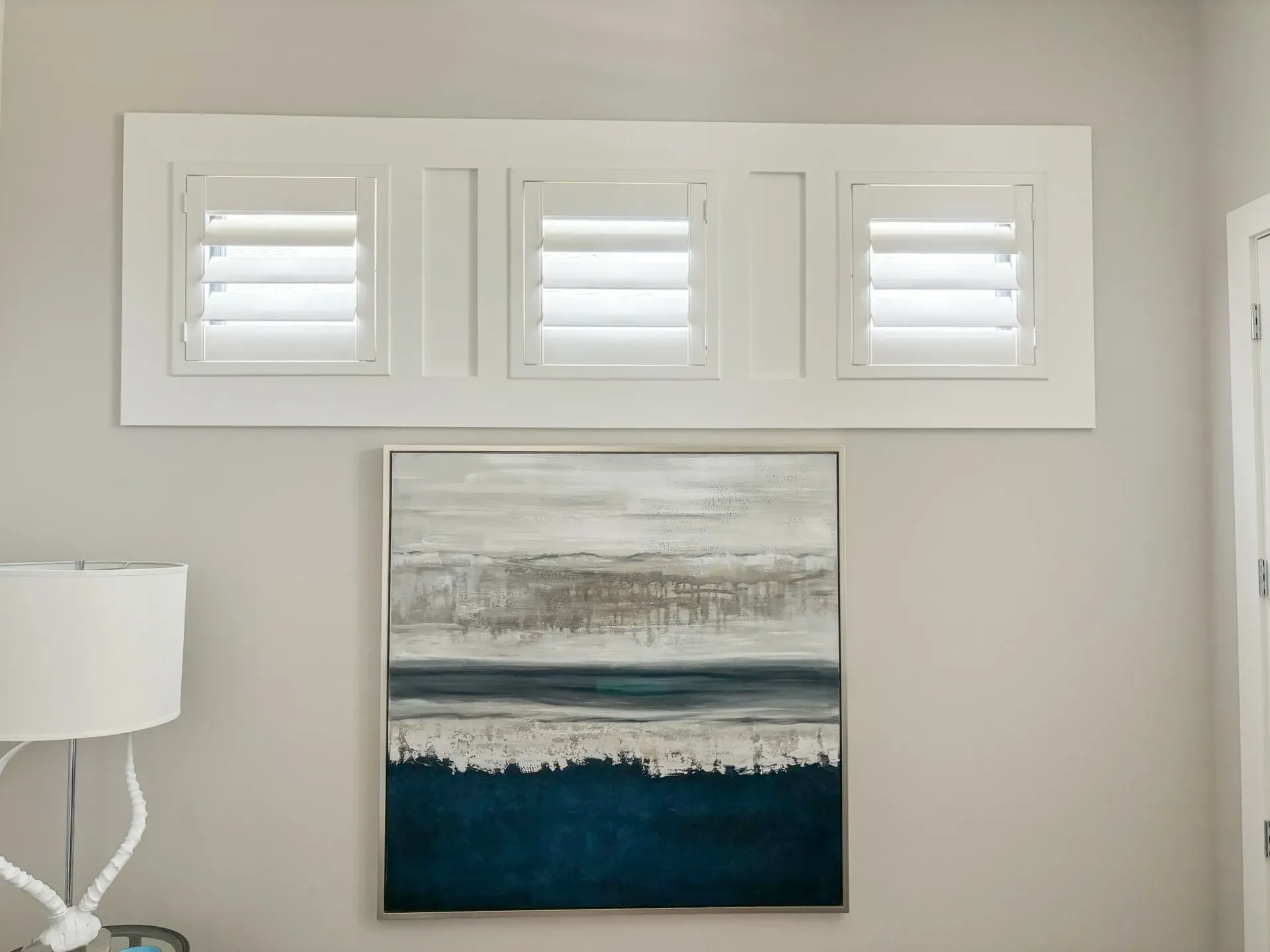 White shutters above an abstract painting on a pale gray wall. A lamp is to the left.