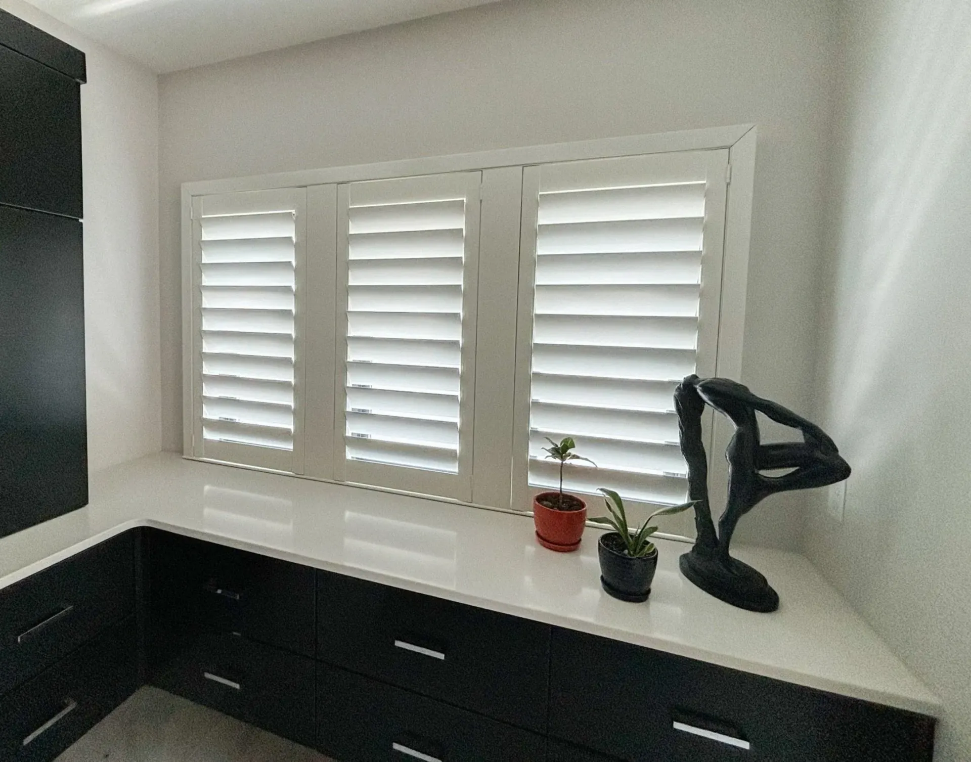 White shuttered window above a countertop with black cabinets, two small plants, and a black sculpture.