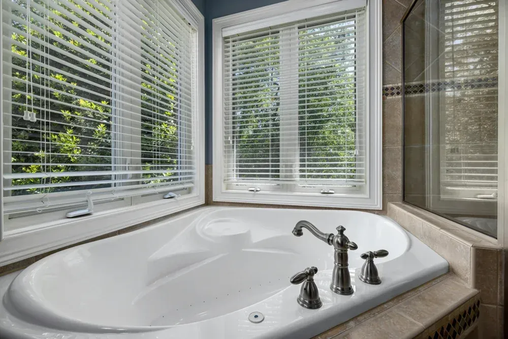 Corner bathtub with blinds, in front of windows with a view of trees.