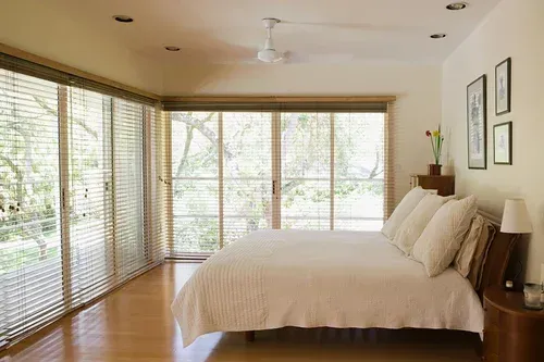 Bedroom with large windows, bed, nightstand, and art on the wall. Natural light streams in.