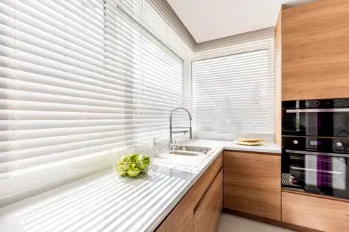Kitchen corner with white blinds, sink, wooden cabinets, and a head of lettuce on the countertop.