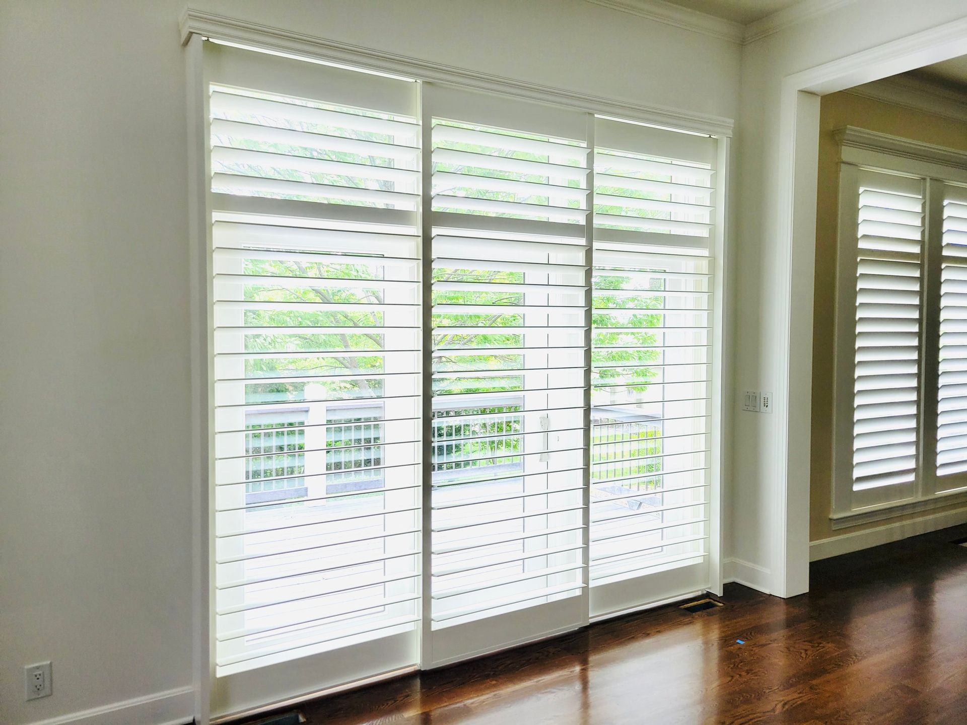 White interior shutters on three sliding glass doors, looking out to a balcony and greenery.