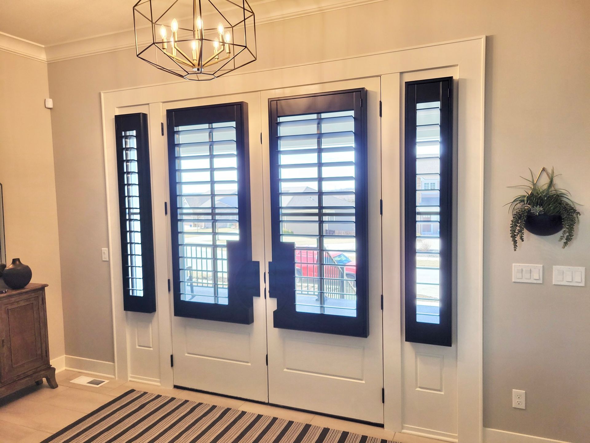 Entryway with black louvered shutters on white doors and sidelights, overhead light, gray walls, and rug.