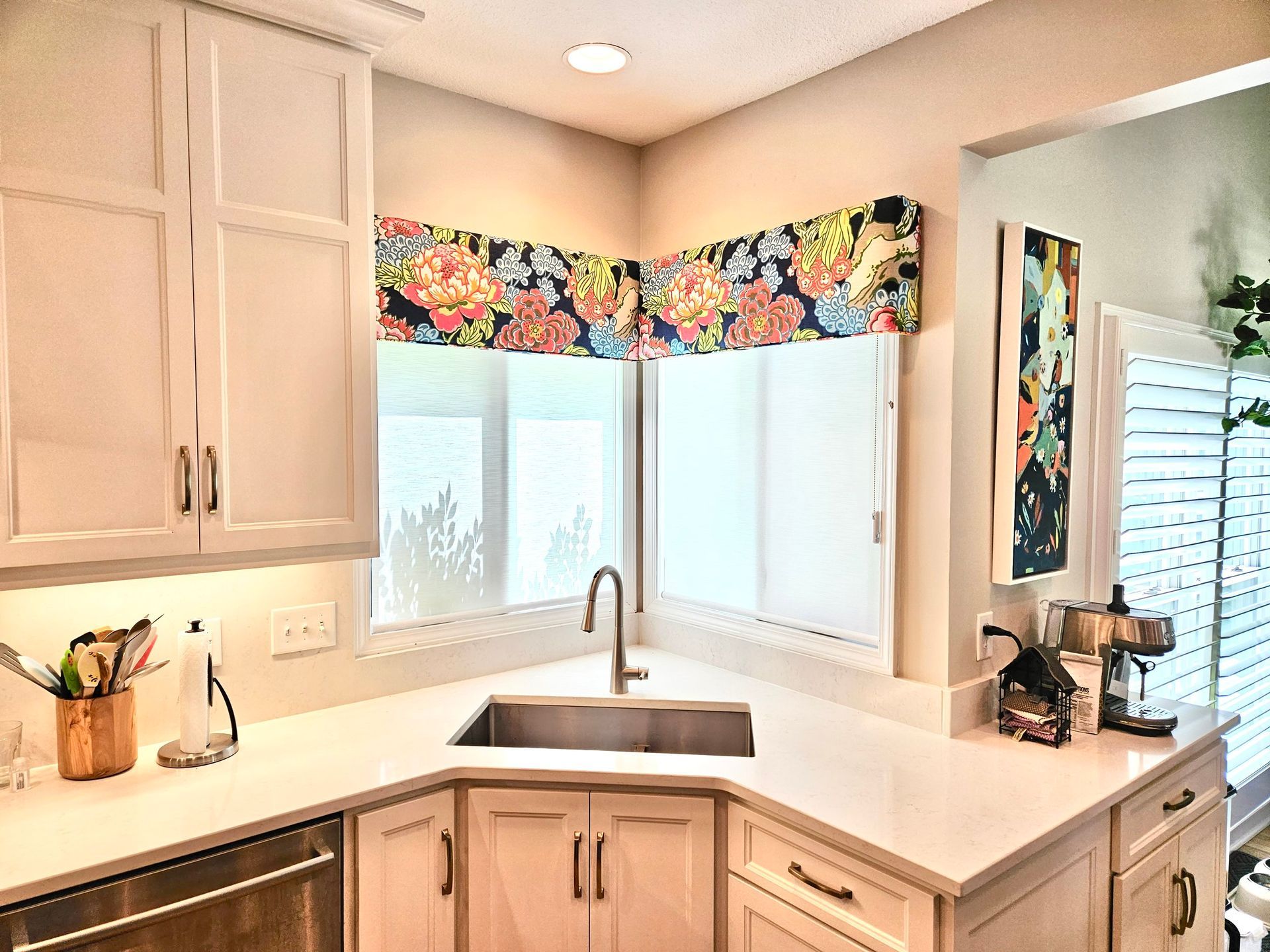 Corner kitchen with white cabinets, countertop, and sink. Floral valance above windows.