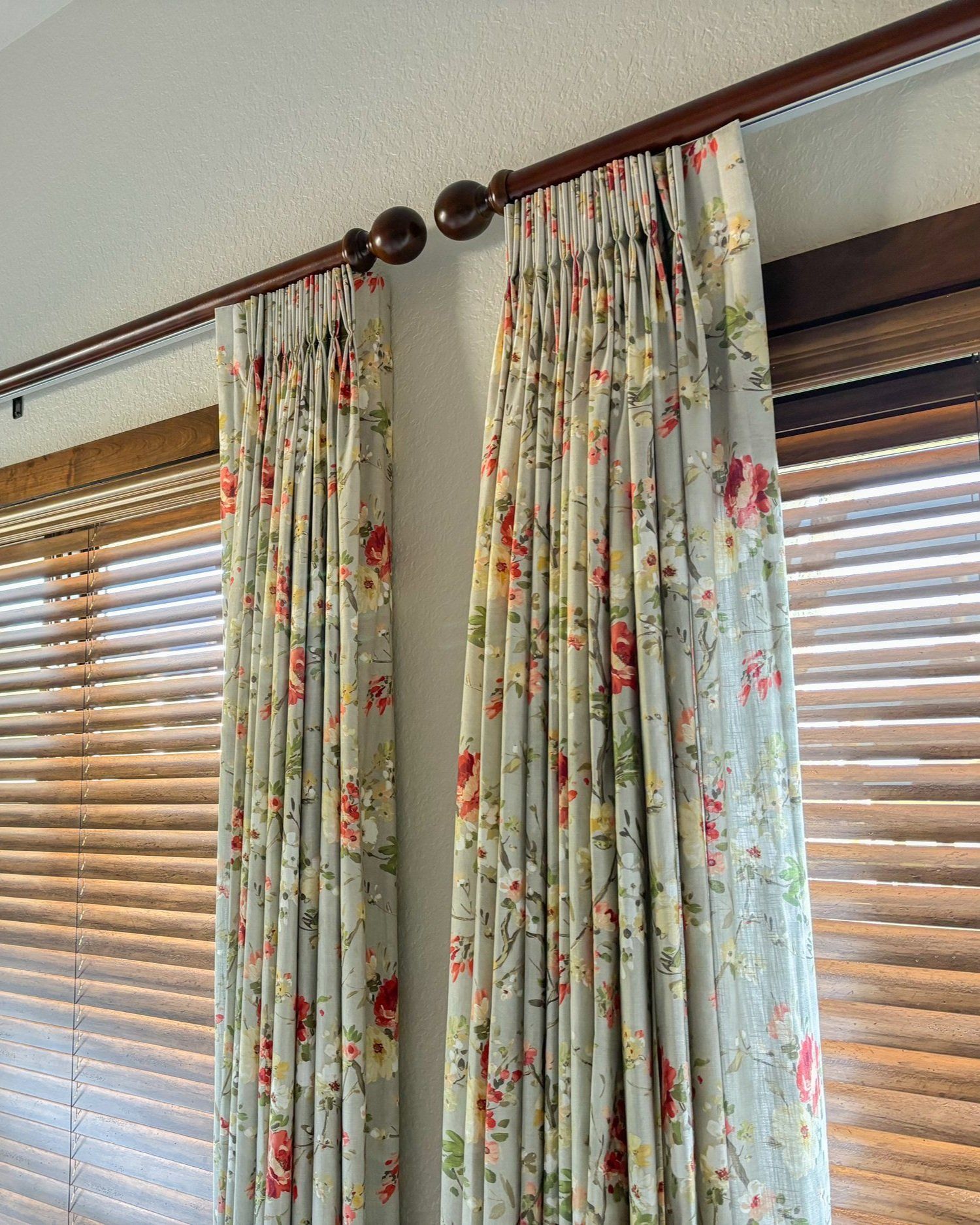 Two floral curtains hanging from a brown rod, in front of wooden blinds.