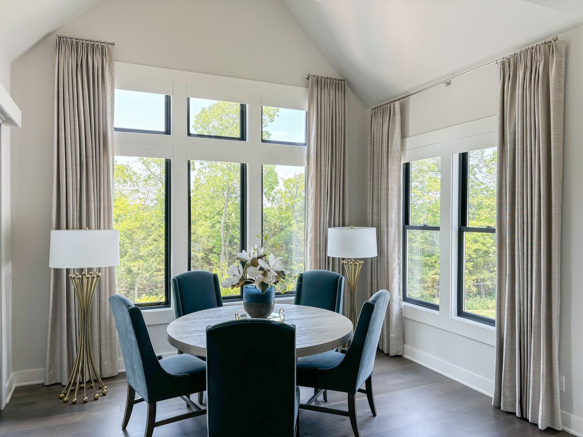 Dining room with a round table, blue chairs, large windows, and beige curtains.