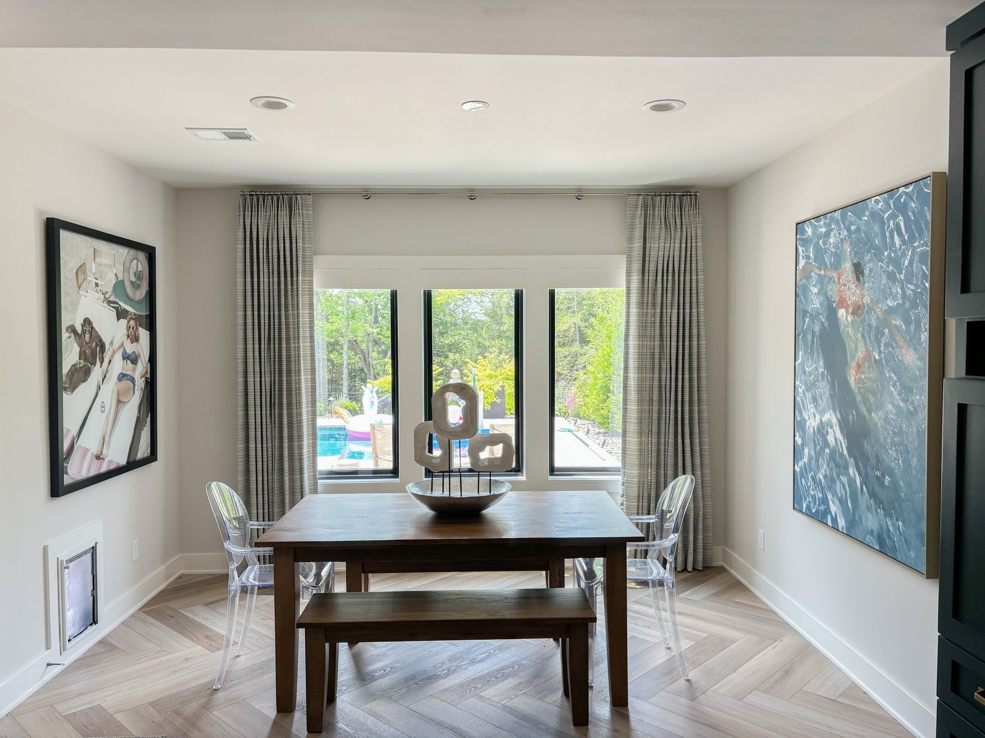 Dining room with wooden table, bench, clear chairs, art, patterned curtains, and a view to a pool.