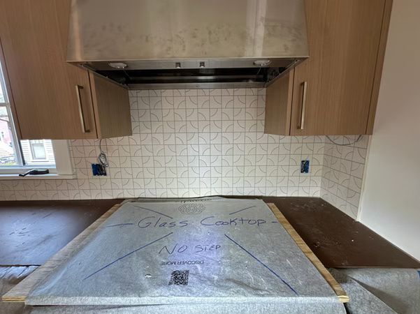 Kitchen under renovation with protected countertop, wood cabinets, white tile backsplash, and range hood.