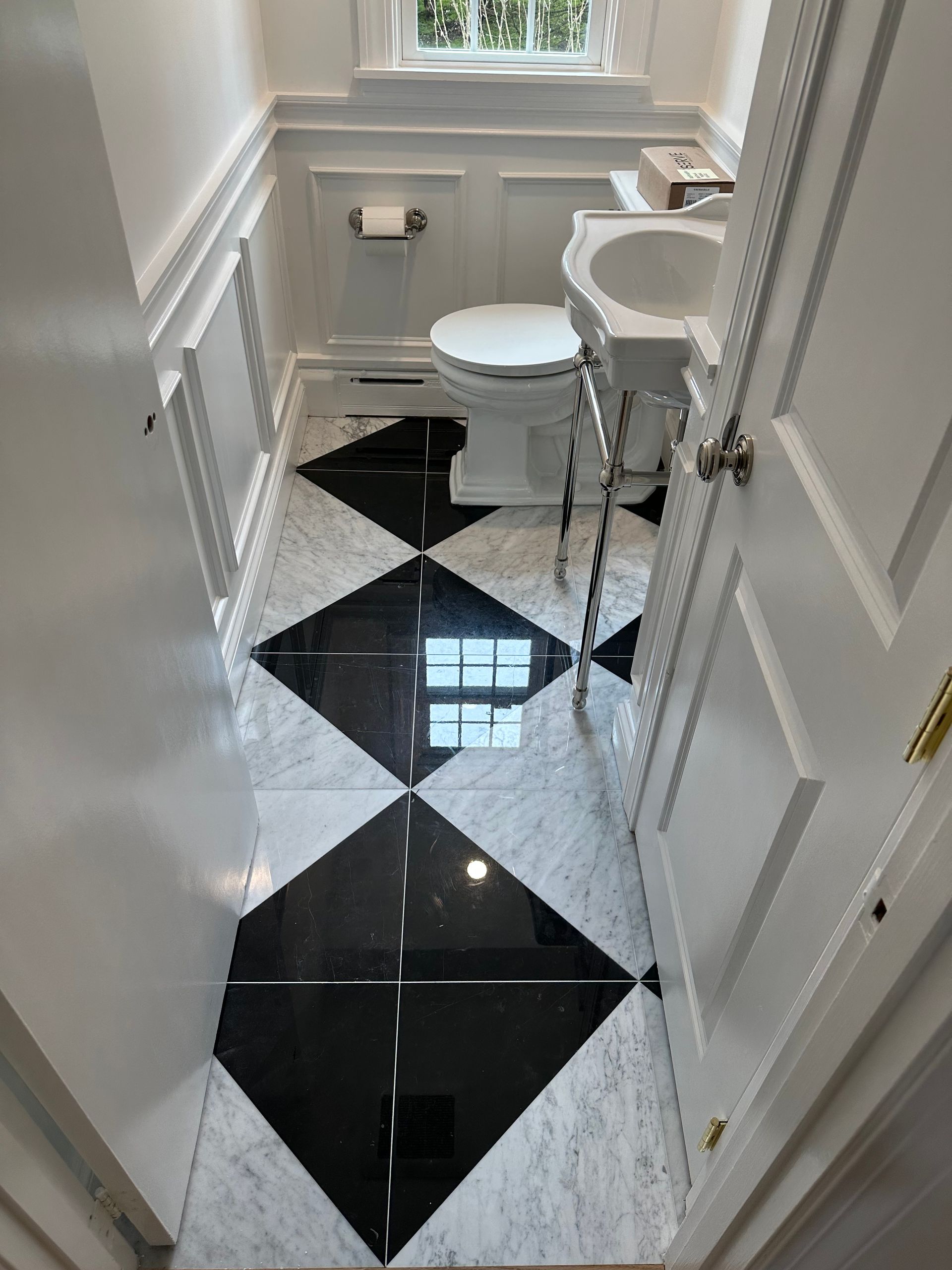 Narrow bathroom with black-and-white diamond tile floor, white sink, toilet, and window.