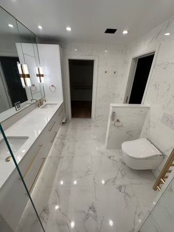 Modern white marble bathroom with glass shower, double vanity, and toilet under bright recessed lights