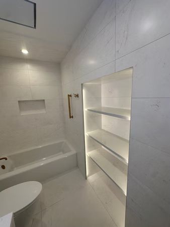Modern white bathroom with built-in shelving, bathtub, and toilet, lit by a wall light.