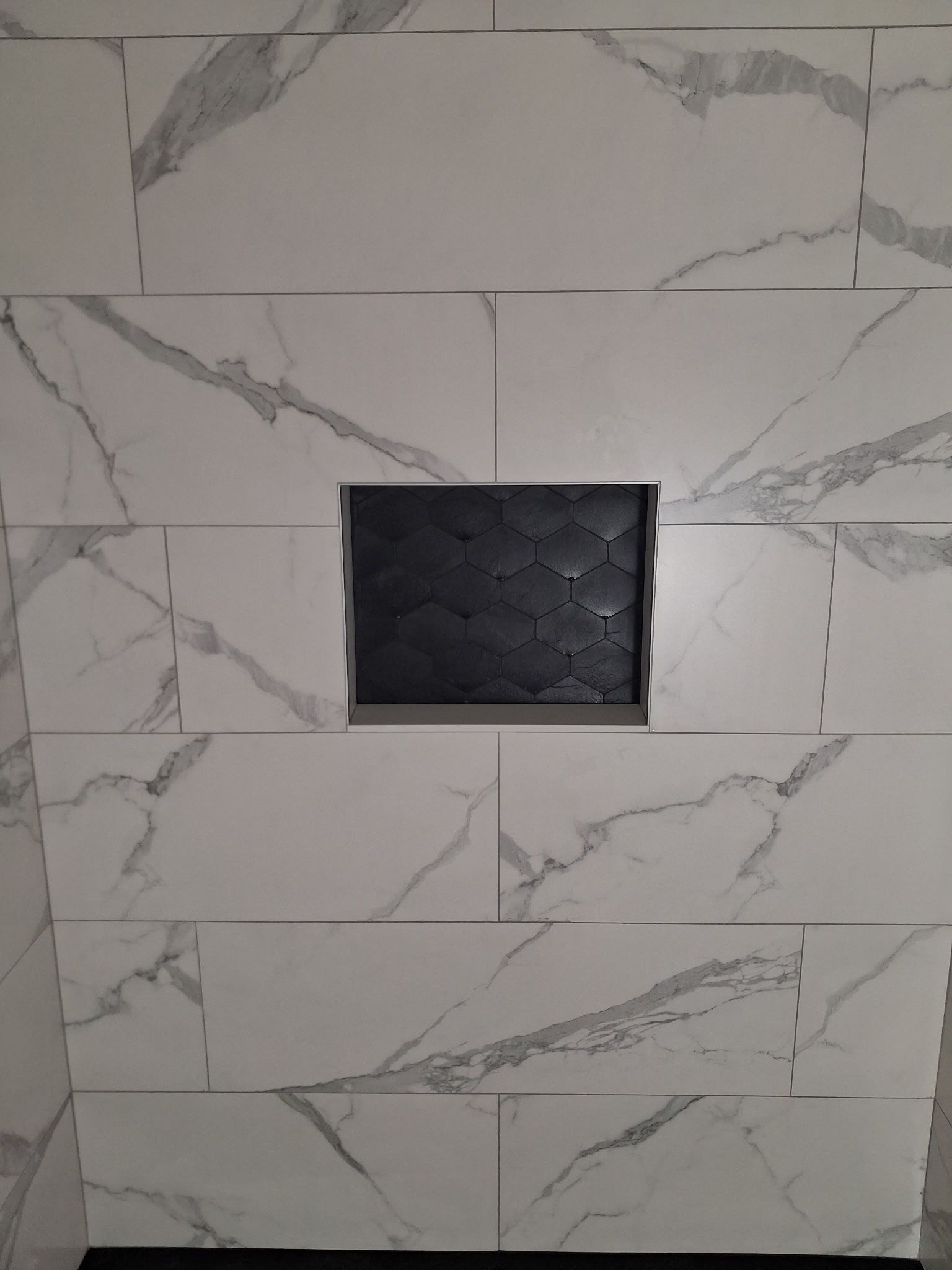 White marble-look tiled wall with a small black square recessed opening in the center.
