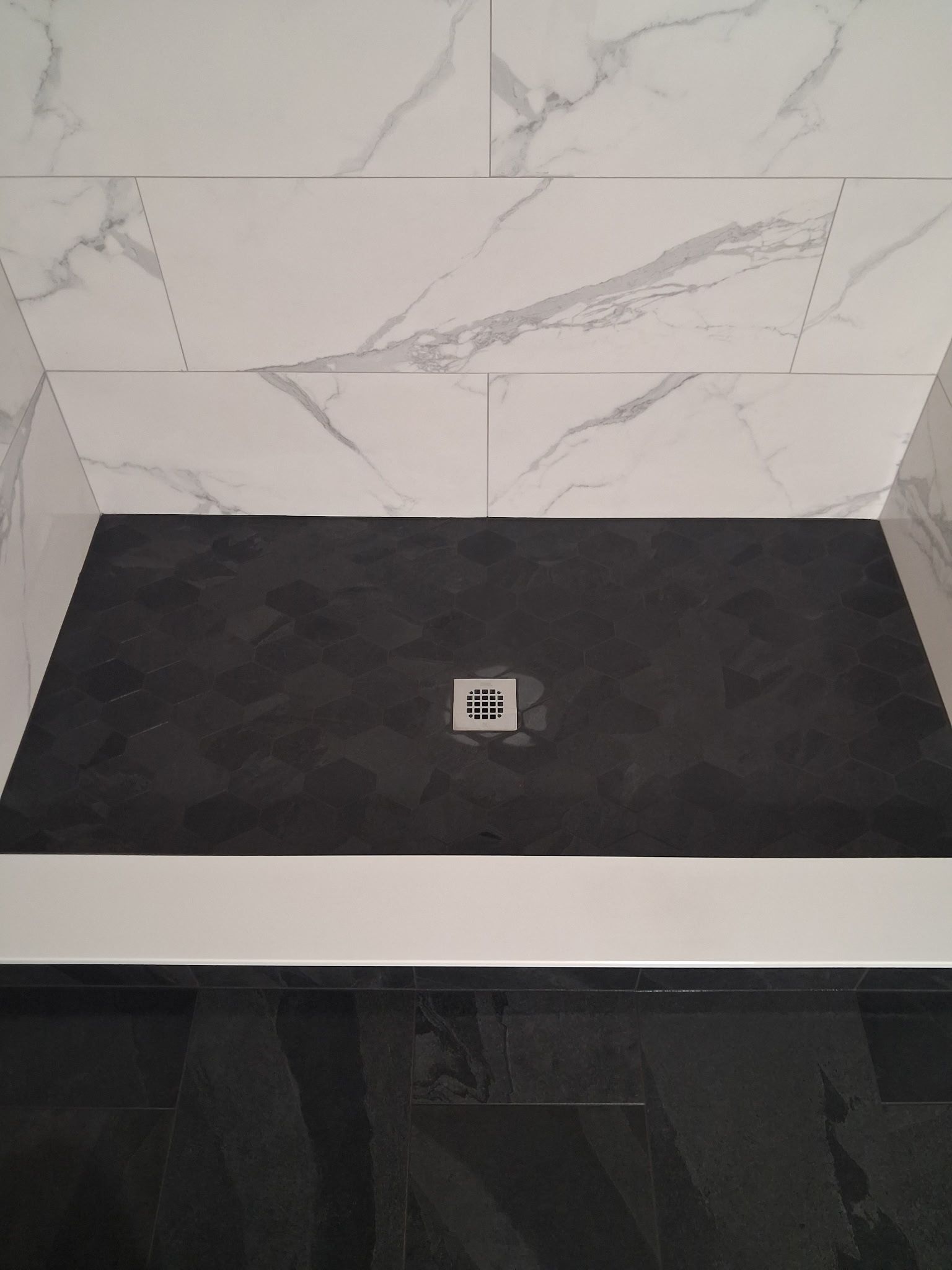Empty shower base with black floor, white marble-pattern wall tile, and a centered drain.
