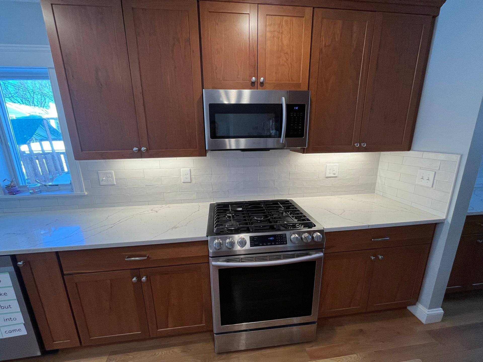 Kitchen with wood cabinets, white counters, stainless steel microwave, and gas range under bright backsplash lighting