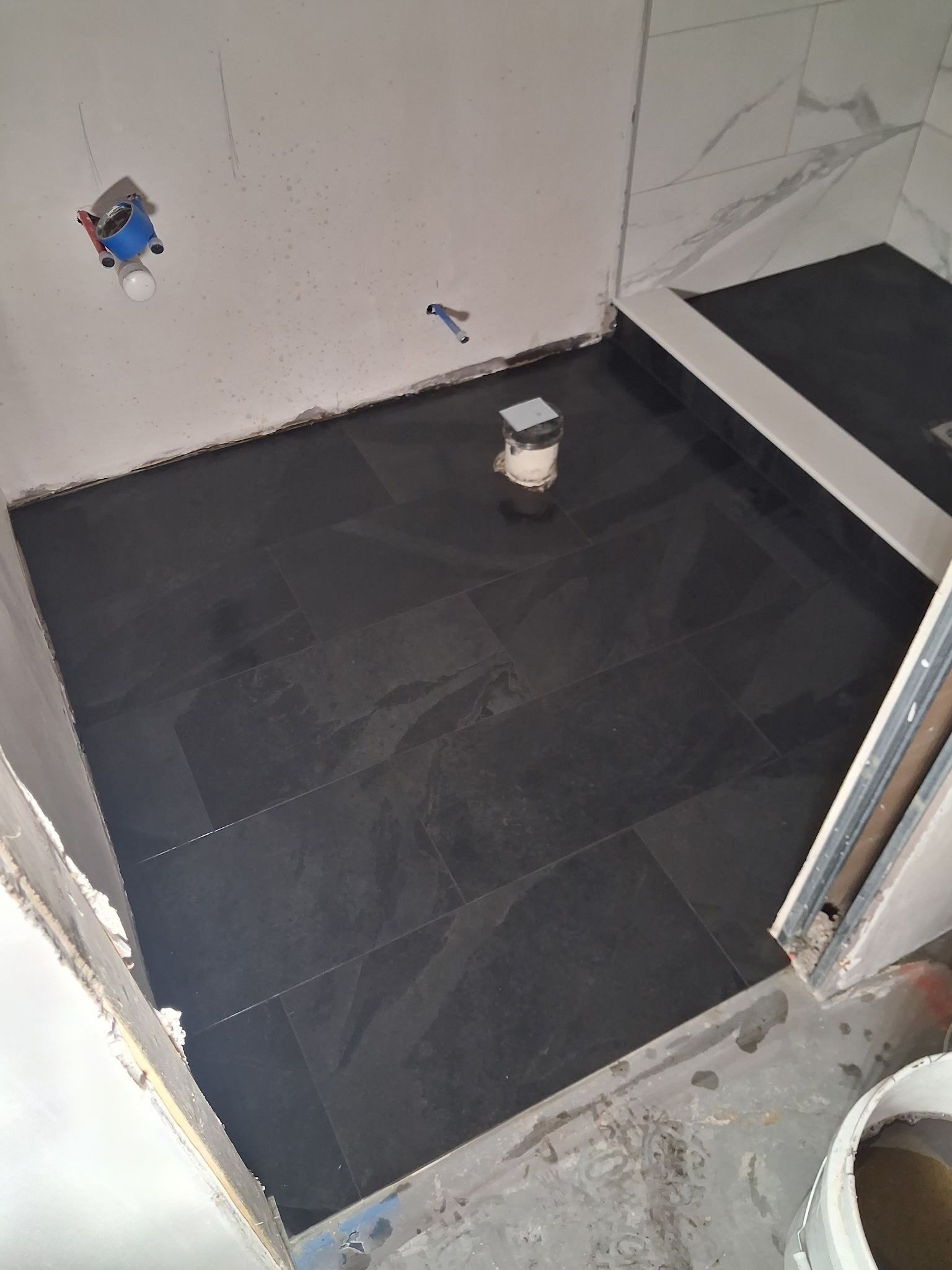 Small tiled room under renovation with black floor tiles, exposed plumbing, and a step in the corner.