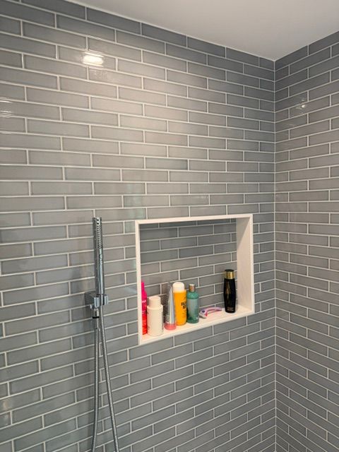 Gray tiled shower with recessed shelf holding toiletries and a handheld showerhead on the wall.
