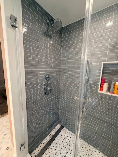 Modern walk-in shower with gray tile walls, patterned floor, glass door, and recessed shelf of toiletries.