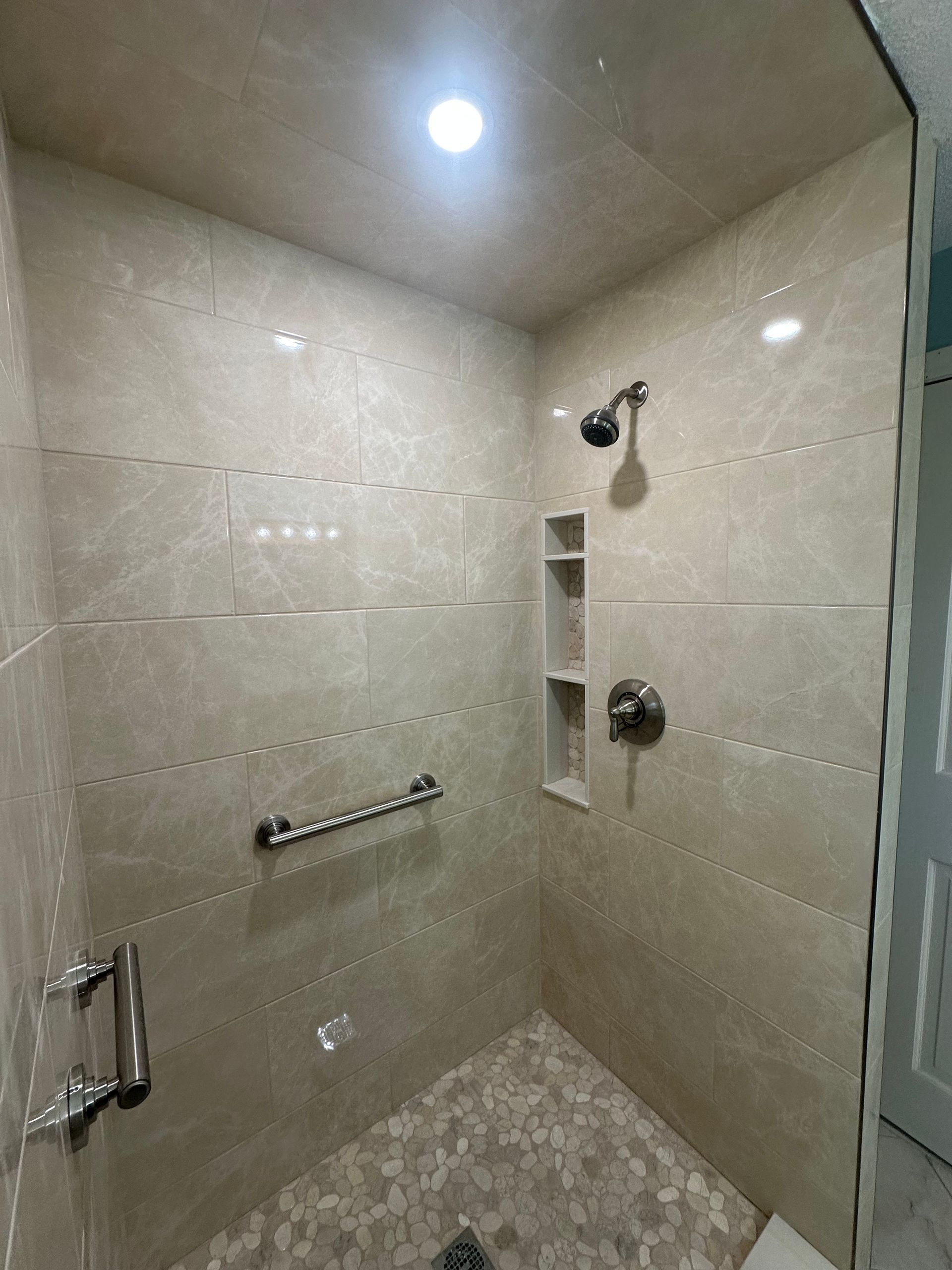 Modern tiled shower stall with wall niche, grab bar, showerhead, and bright ceiling light