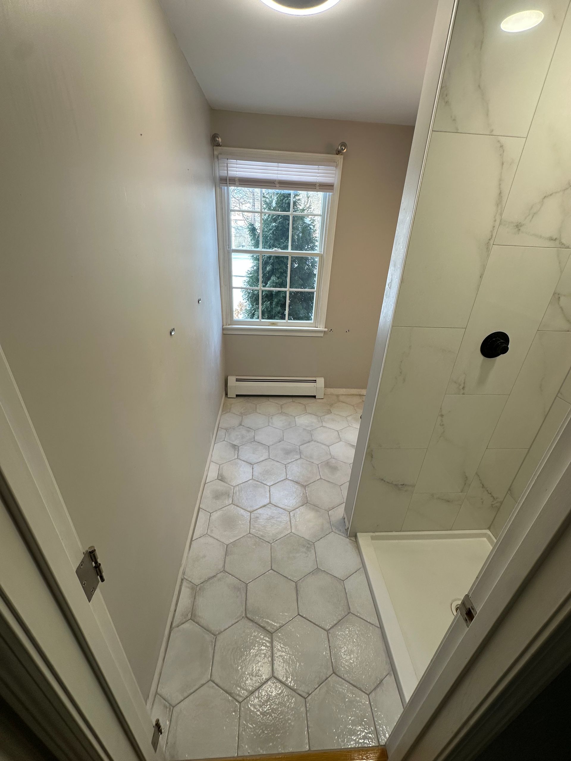 Narrow bathroom with white hex tile floor, shower stall on right, and window at the far end.