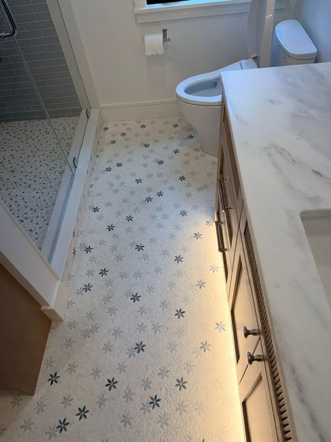 Narrow bathroom with walk-in shower, toilet, vanity, and light patterned floor tiles.