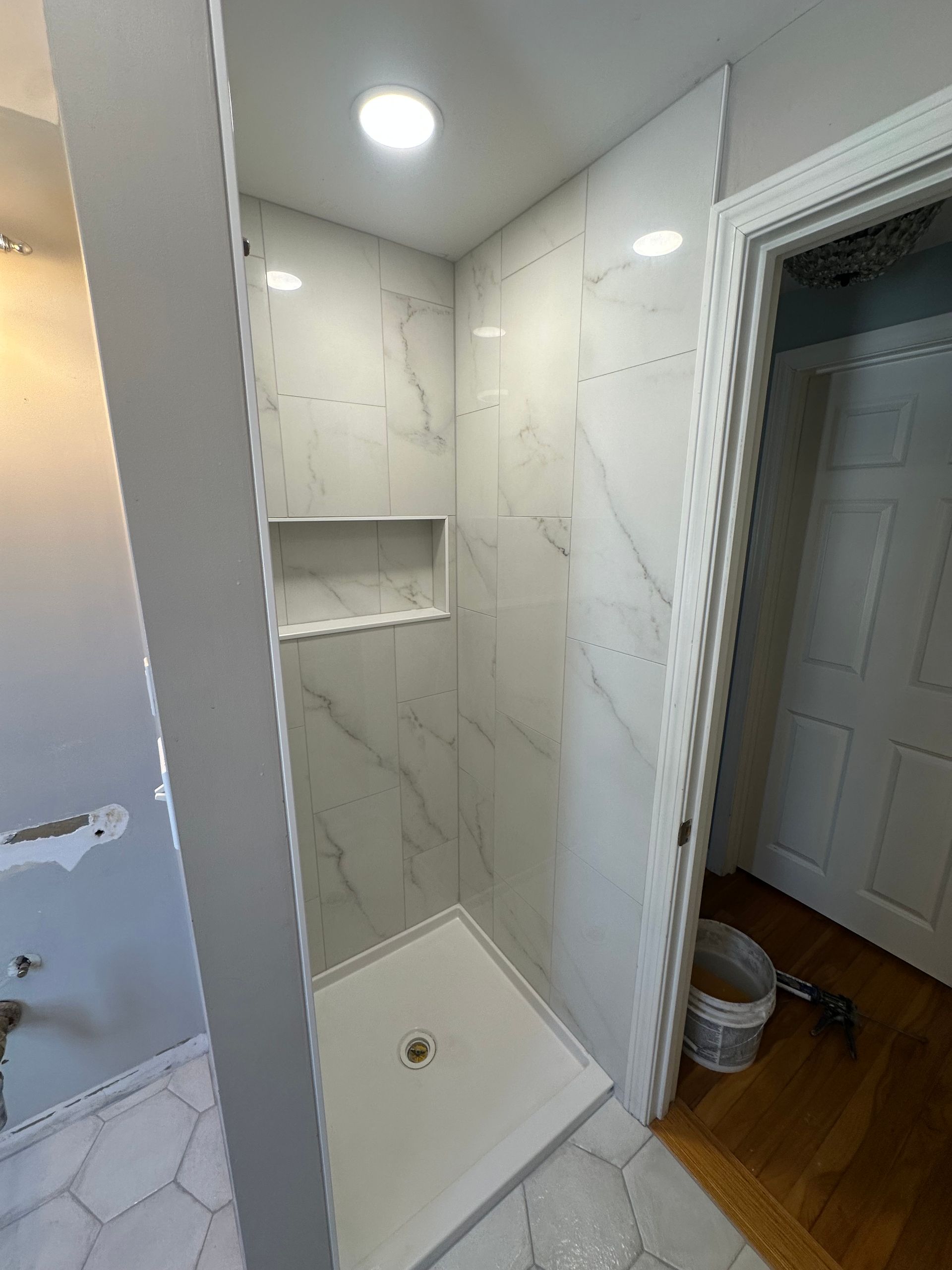 Small tiled shower stall with built-in shelf, white pan, and an open door beside wood flooring.