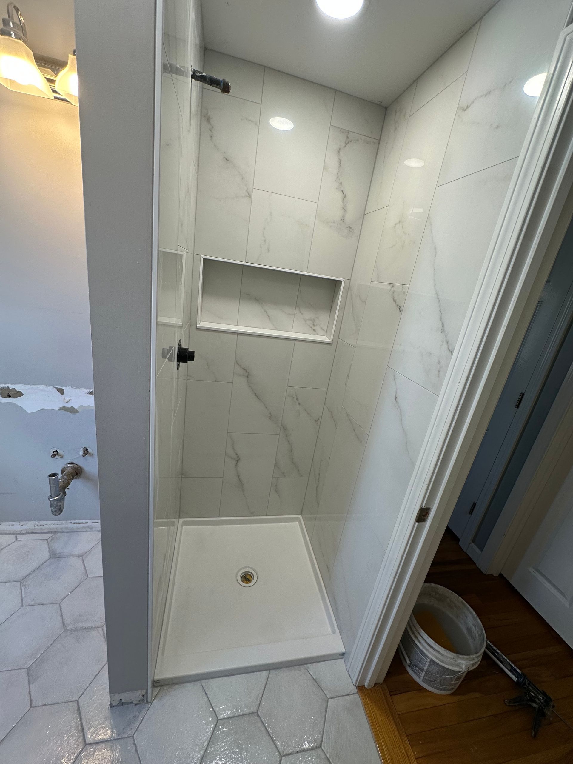 Small tiled walk-in shower with white marble-look walls, shelf, and bench seat near a doorway.