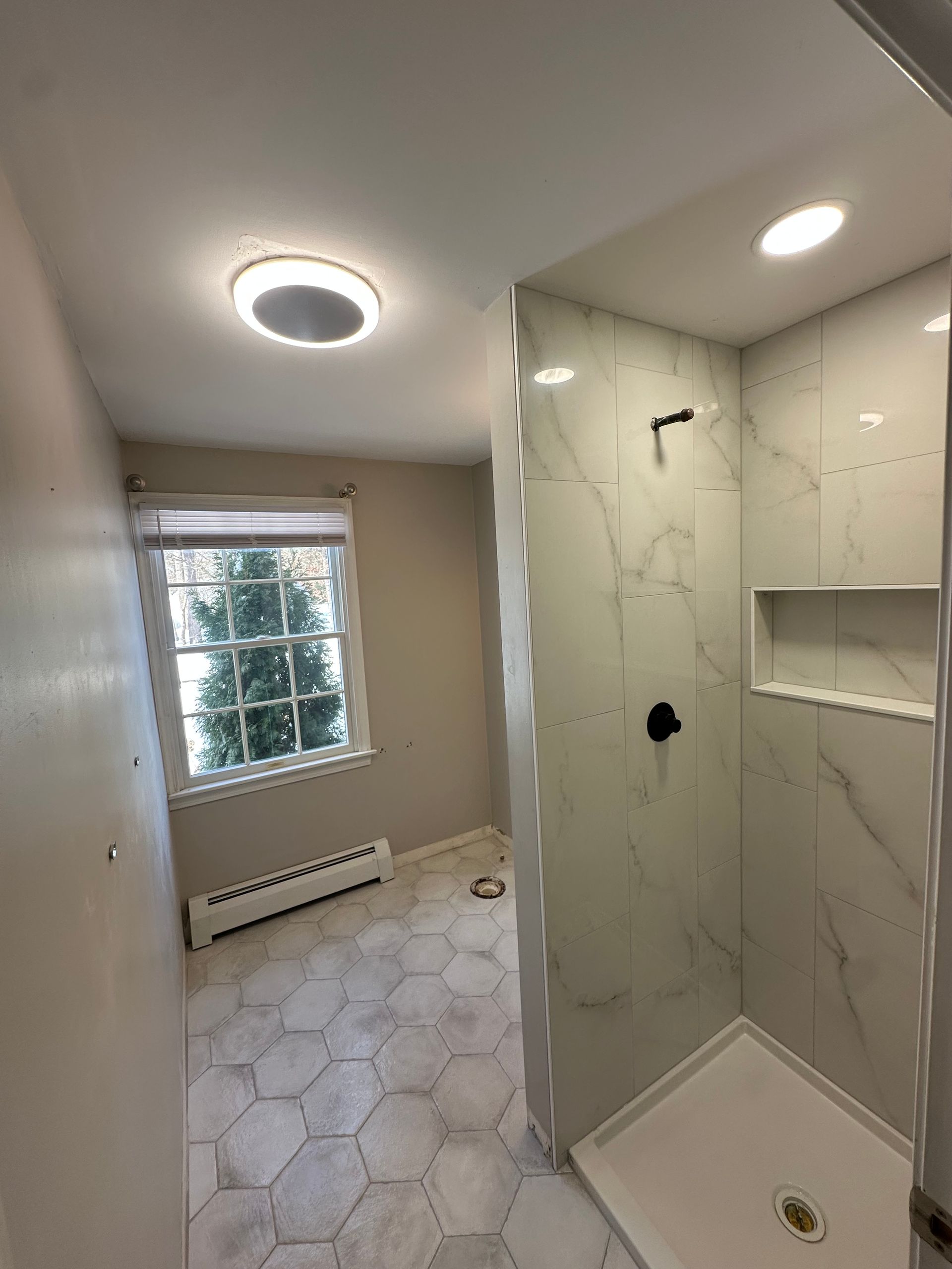 Small bathroom with walk-in shower, hex tile floor, marble-look walls, and a window with radiator beneath.