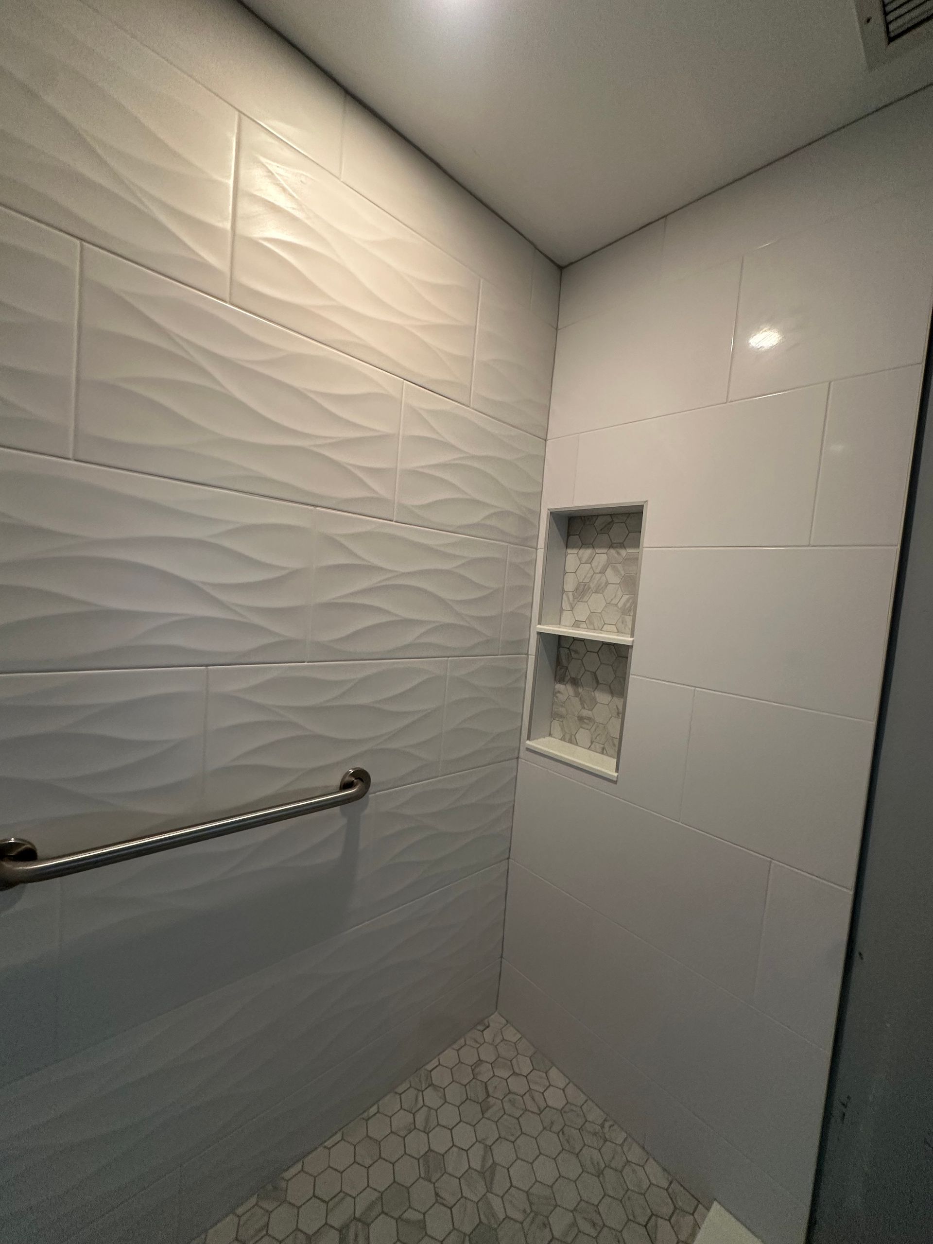 White tiled shower with textured accent wall, grab bar, and recessed shelf niche.