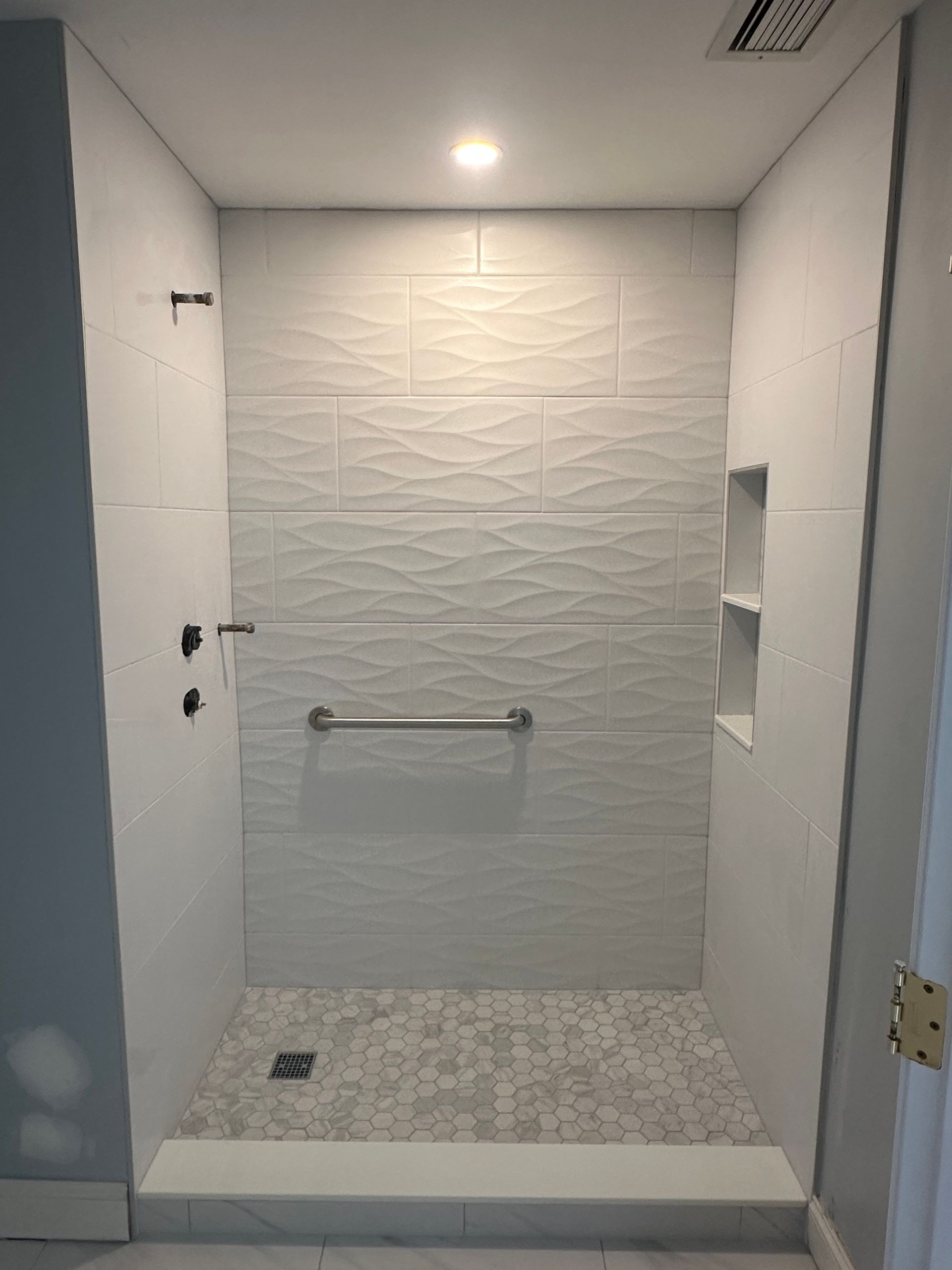 Empty accessible shower stall with white tile walls, grab bar, and mosaic floor.