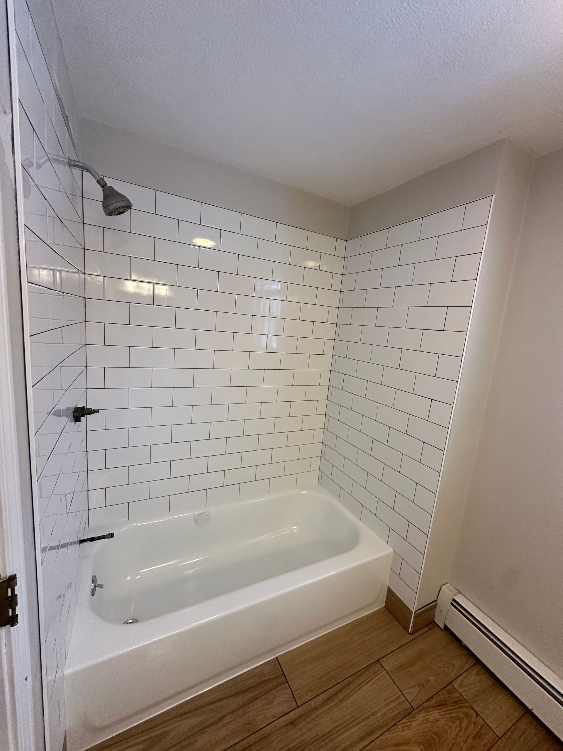 Small white bathroom with a bathtub, tiled shower walls, and wood-look flooring.