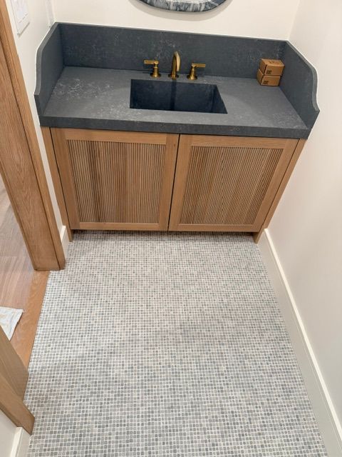 Small bathroom vanity with sink, gold faucet, and wood cabinet on a light patterned floor.
