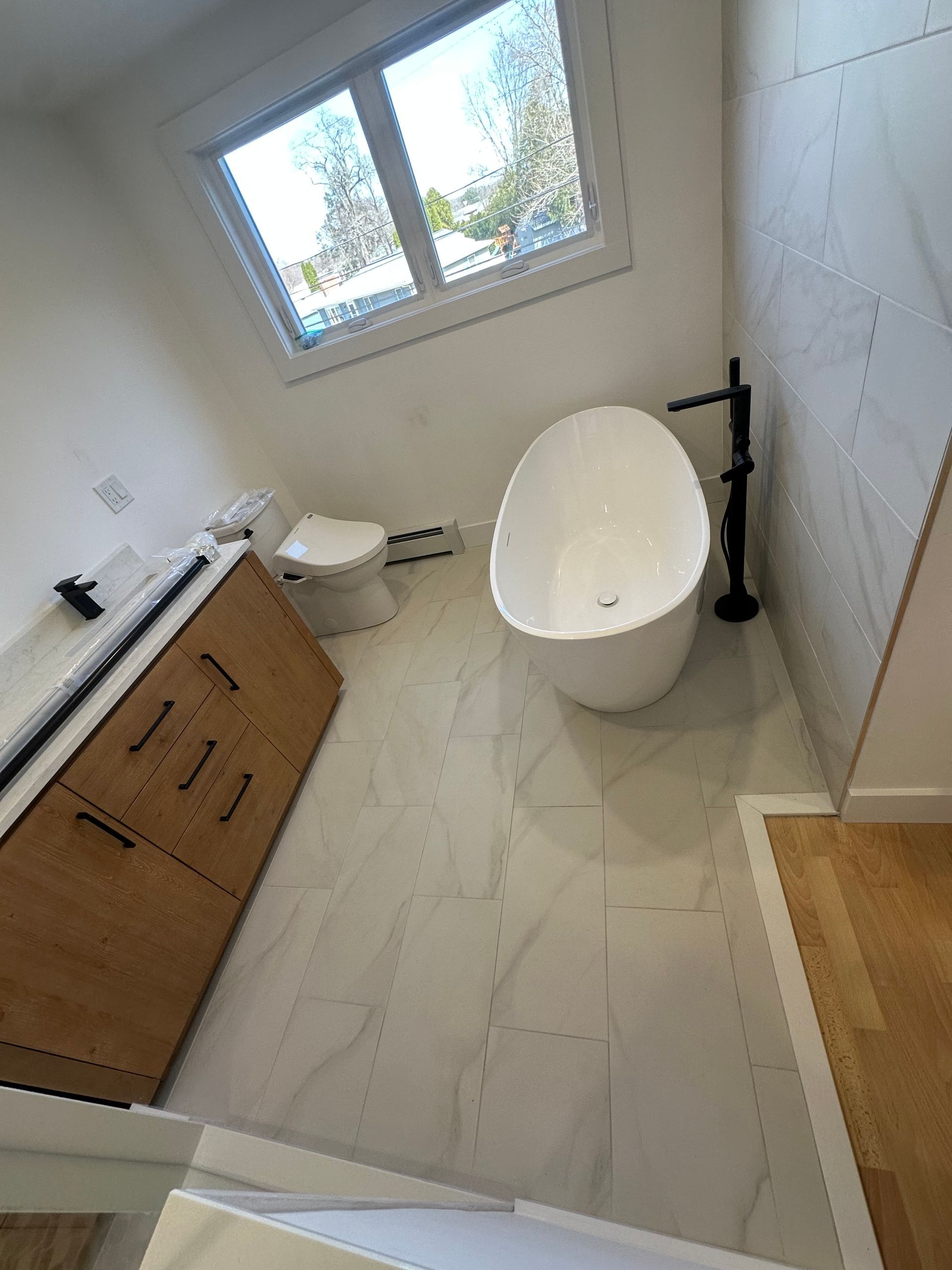 Modern bathroom with freestanding tub, toilet, wood vanity, and tiled floor by a window