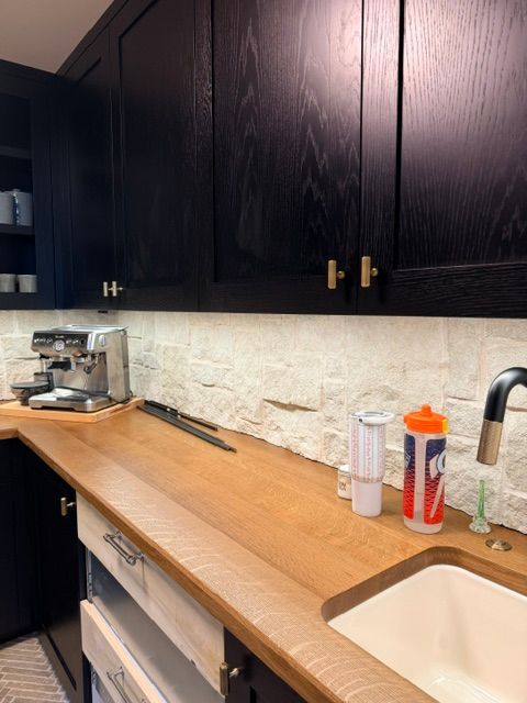 Kitchen counter with dark cabinets, white backsplash, wooden countertop, espresso machine, sink, and orange bottle organizer.