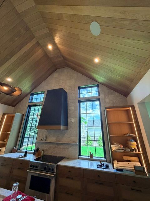 Modern kitchen with wood ceiling, black stove hood, and tall windows overlooking greenery.