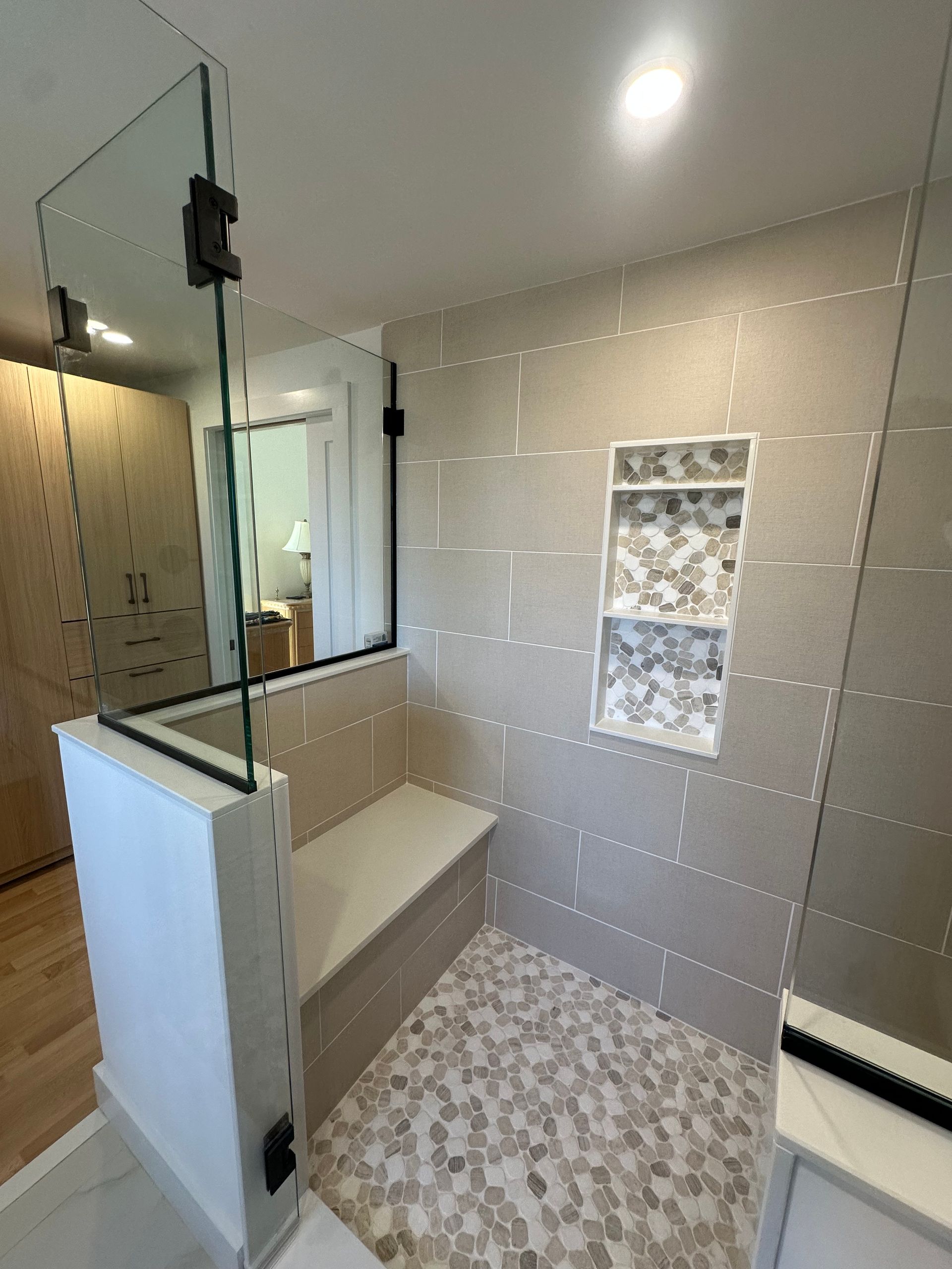 Modern bathroom with glass shower, white bench, beige tiled walls, and pebble-style floor tiles