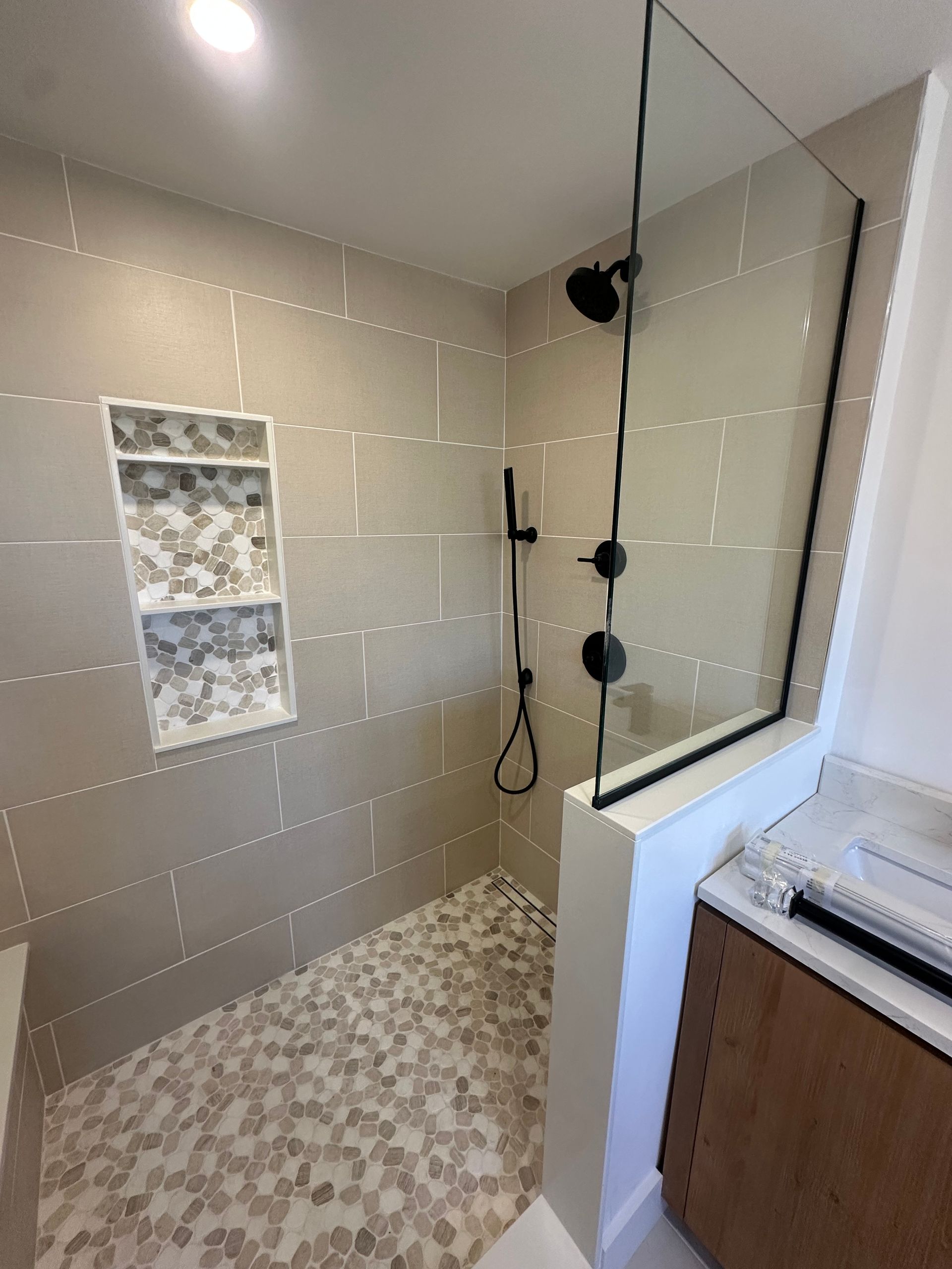 Bathroom shower with beige tile, pebble floor, black fixtures, and a glass partition beside a vanity.