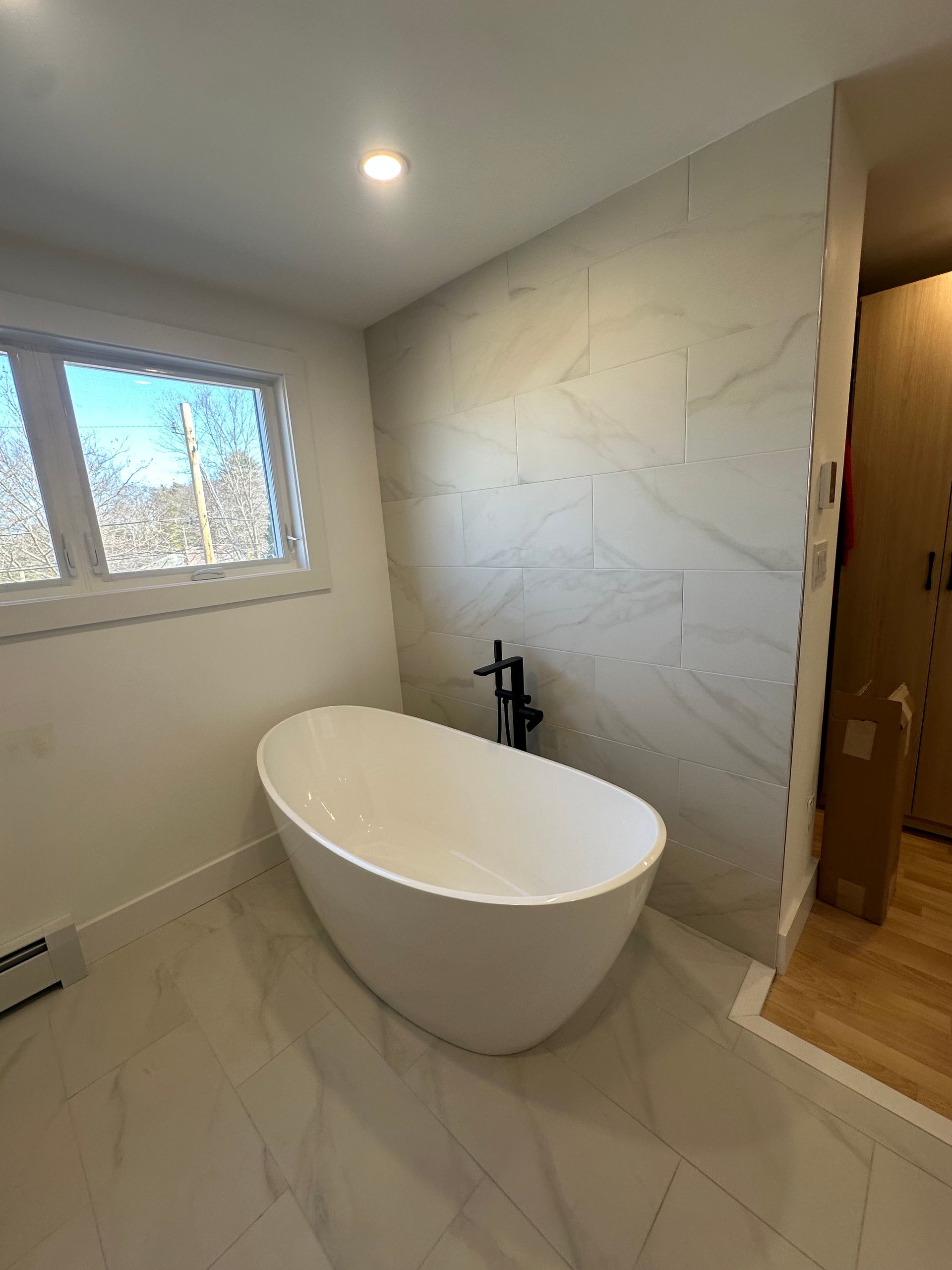 Modern bathroom with a freestanding oval tub, tiled walls, window, and wooden floor entryway.