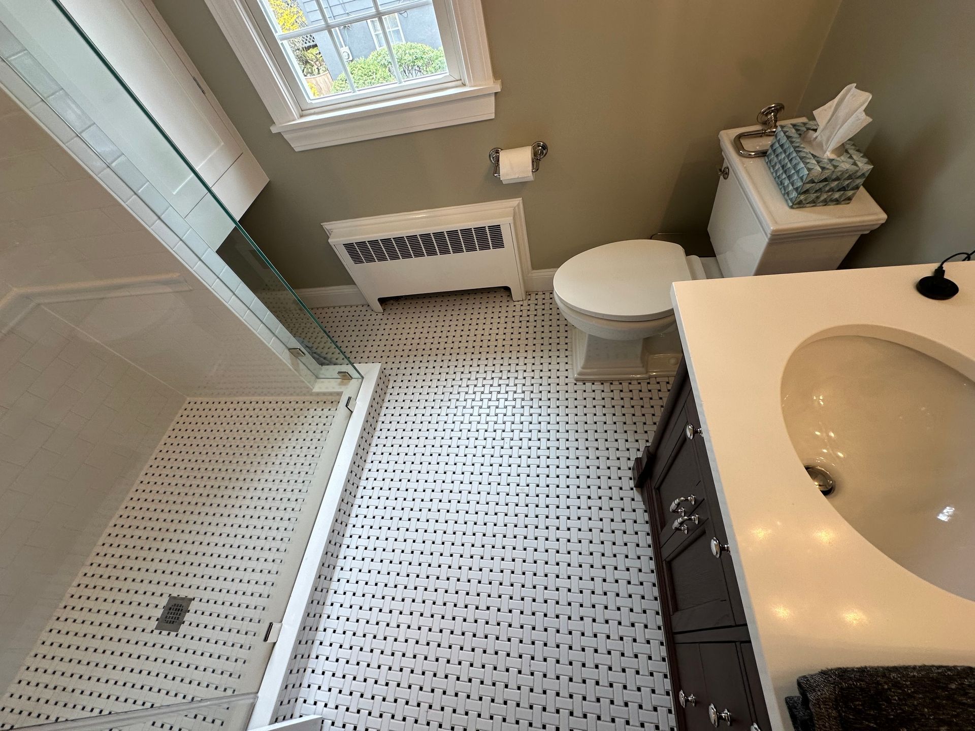 Small bathroom with white tile floor, glass shower, toilet, sink vanity, and window above a radiator.