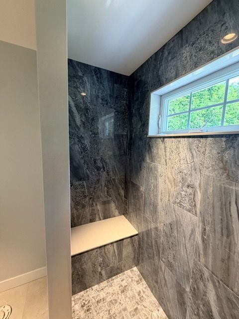 Narrow walk-in shower with dark gray marble walls, white bench, and a high window letting in daylight
