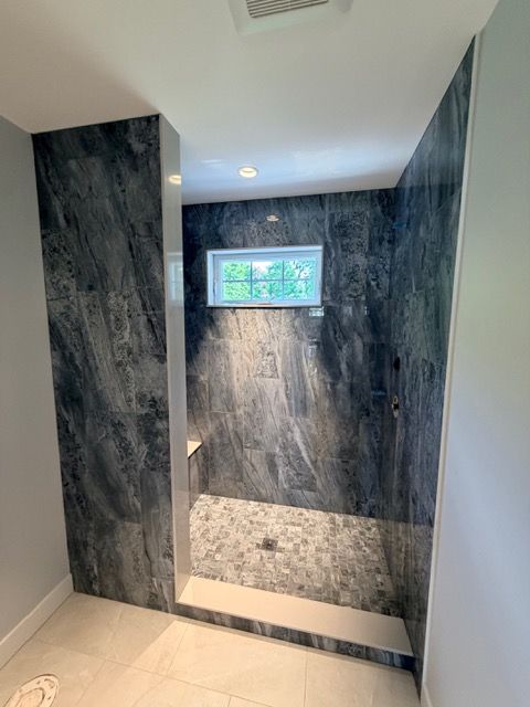 Modern bathroom shower with dark stone walls, white tile floor, and a small window.
