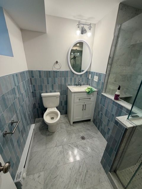 Narrow bathroom with blue tile, toilet, vanity sink, oval mirror, and glass shower stall.