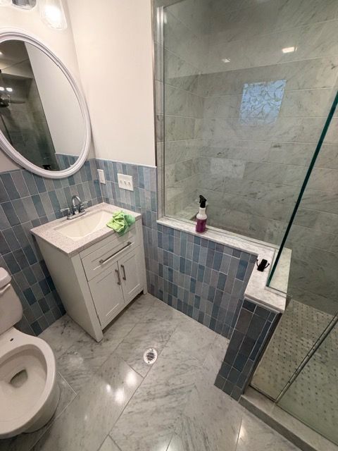 Small tiled bathroom with sink vanity, toilet, glass shower, and gray-blue marble-pattern walls and floor.