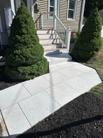 Front walkway with light stone pavers leading to a house entrance, flanked by two trimmed evergreen shrubs.