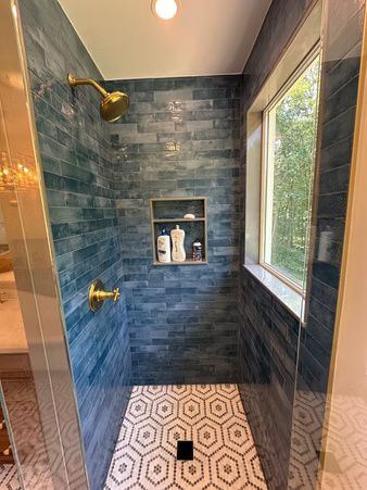 Blue-tiled walk-in shower with brass fixtures, patterned floor, and a window letting in natural light