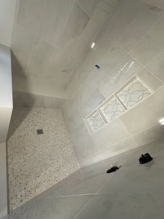 Tilted view of a modern tiled bathroom floor and wall with a shower drain and two black sandals