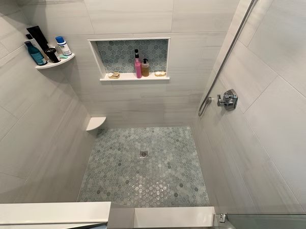 Small tiled shower with gray floor, built-in shelf, window, and soap dispenser on the wall.
