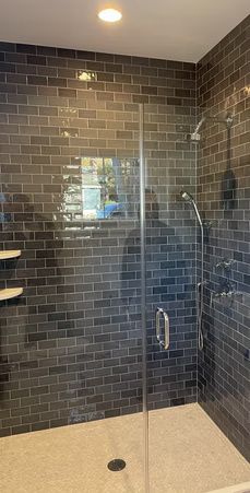 Modern shower with dark tile walls, clear glass door, and beige stone floor.