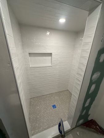 Small tiled shower stall with a recessed shelf, white walls, and a blue floor drain cap